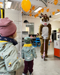 Osterhase fährt auf einem Roller durch den KS Ausstellungsraum und Kinder schauen begeistert zu