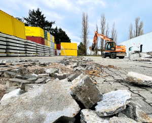 Im Vordergrund Trümmer von Beton und Pflastersteinen, während im Hintergrund ein oranger Mobilbagger vor gestapelten Containern und Betonfertigteilen auf einem Betriebsgelände arbeitet