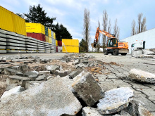 Im Vordergrund Trümmer von Beton und Pflastersteinen, während im Hintergrund ein oranger Mobilbagger vor gestapelten Containern und Betonfertigteilen auf einem Betriebsgelände arbeitet